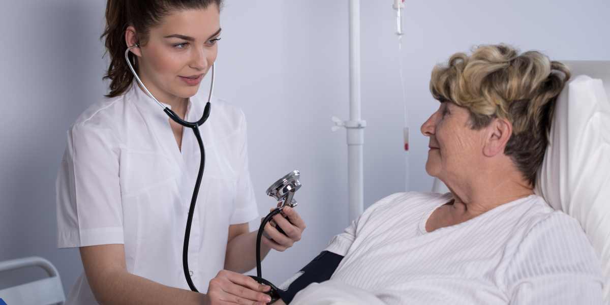 A young female nurse in a white uniform checks the blood pressure of an elderly female patient resting in a hospital bed, using a stethoscope and a manual blood pressure cuff.